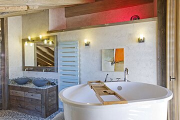 A modern rustic bathroom features a white freestanding tub with a caddy, a double sink vanity, abstract art, exposed wooden beams, and red accent lighting.