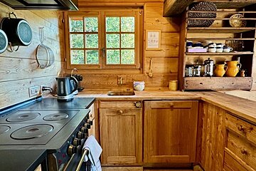 Cozy, rustic wooden kitchen with timber walls, a window over the sink, a black stove, and open shelving filled with mugs and pitchers.