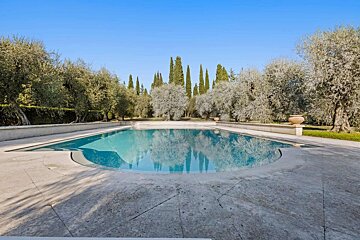 A serene swimming pool reflects cypress and olive trees under a clear blue sky, surrounded by a lush, green landscape.