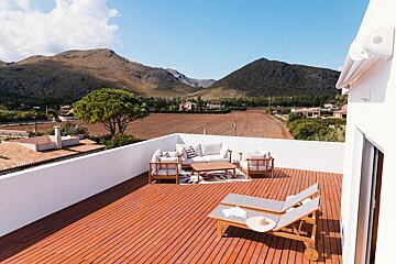 A modern rooftop deck with outdoor seating and a sun lounger offers stunning views of a rural valley, mountains, and fields under a bright blue sky.