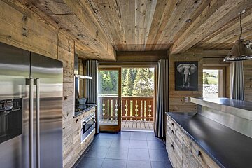 A modern rustic kitchen featuring weathered wood walls/ceiling, dark tiled floor, stainless steel appliances, and a balcony with a forest view.