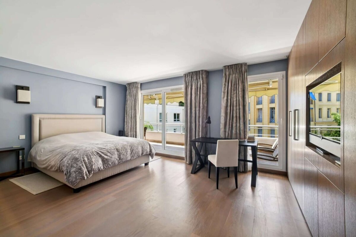 A modern bedroom with light blue walls, a large bed, hardwood floors, balcony access, a desk, and wood paneling with an integrated TV.