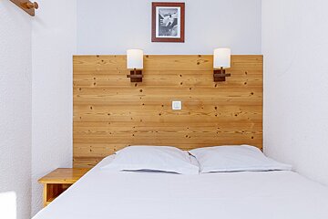 A bedroom with a white bed, wooden headboard, two wall lamps, and a framed picture. A small wooden nightstand is on the left.