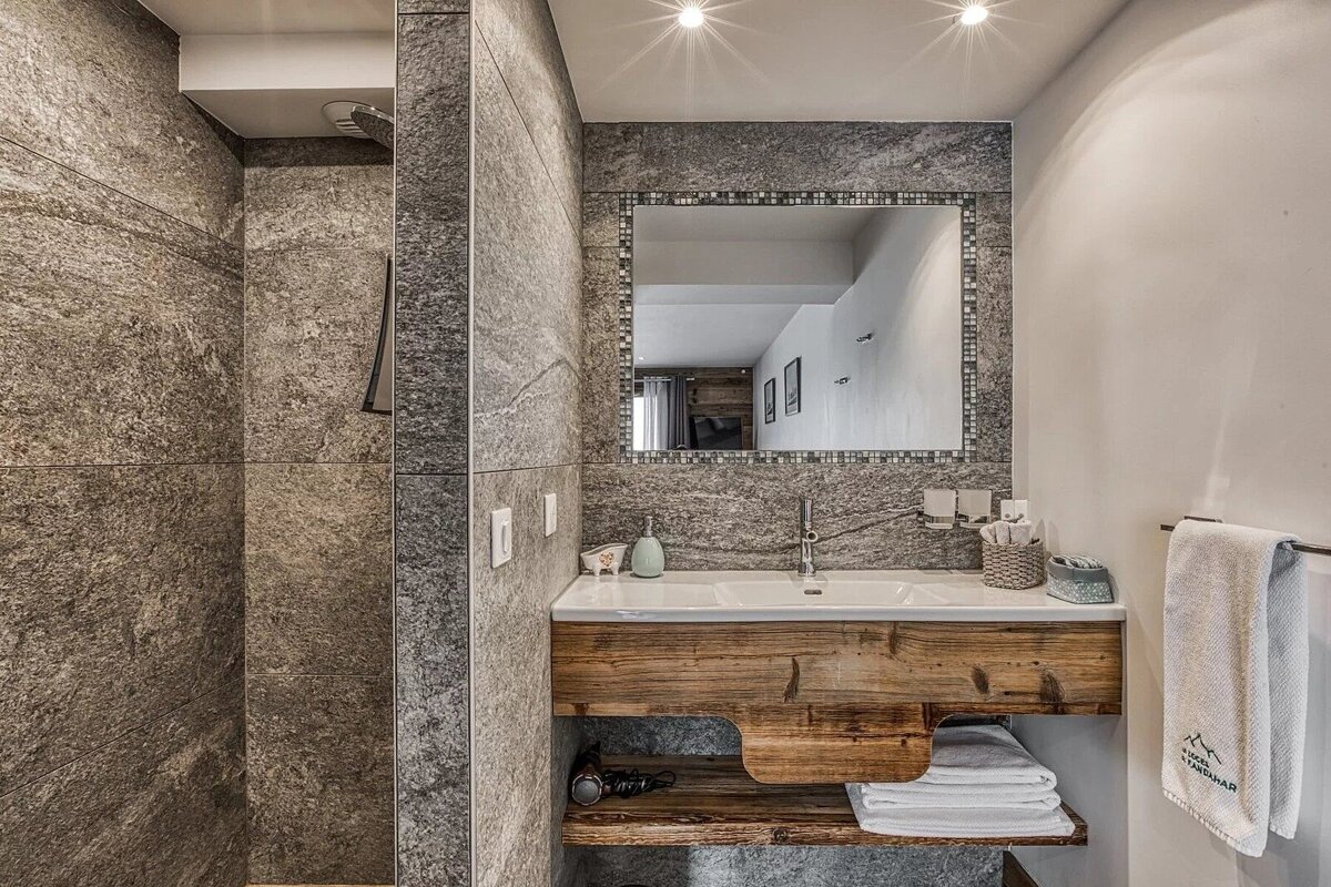 Sleek modern bathroom with grey stone walls, wooden vanity, sink, and mirror. A shower area is visible on the left.