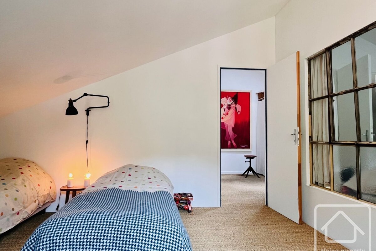A modern, minimalist attic bedroom with two beds. A door opens to a hall featuring a vibrant red painting. Industrial-paneled window on the right.
