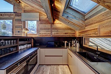 A rustic wooden chalet kitchen with modern beige cabinets, dark counters, a skylight, and snowy mountain views from large windows.