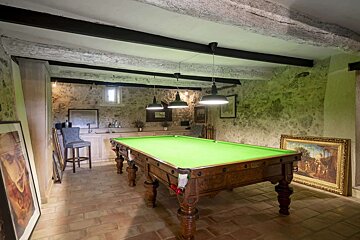 A rustic game room features a vintage snooker table with green felt, stone walls, exposed beams, terracotta floor, and industrial pendant lights.