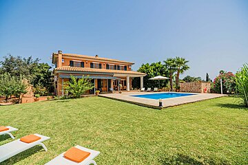 An inviting orange villa features a sparkling blue pool, vast green lawn, and sun loungers under a bright, clear sky, surrounded by lush foliage.