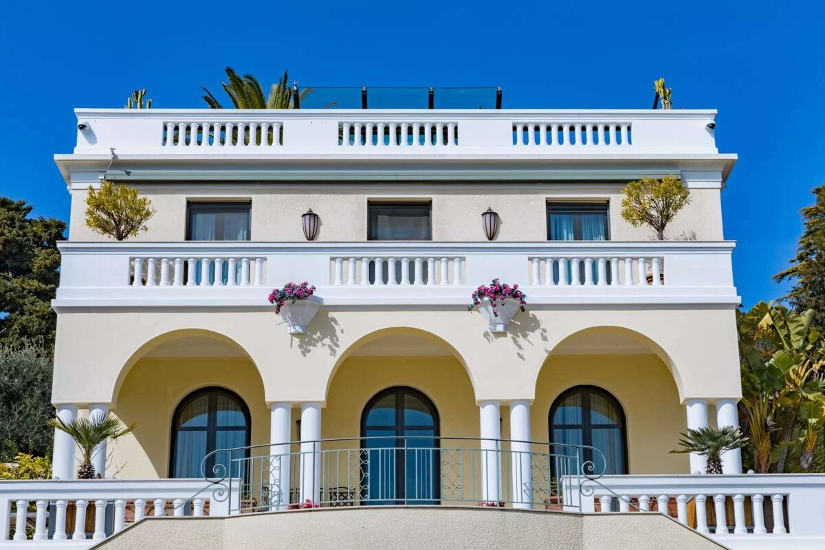 An elegant cream-colored building with multiple balconies featuring white balustrades and arched windows, set against a clear blue sky. Flowering plants adorn the balconies.