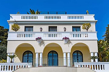 An elegant cream-colored building with multiple balconies featuring white balustrades and arched windows, set against a clear blue sky. Flowering plants adorn the balconies.