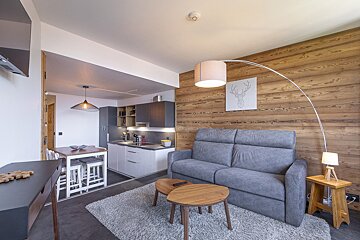 Cozy, modern open-plan studio with a gray sofa, wooden accent wall, and sleek kitchen. A dining table and minimalist decor create a warm, inviting atmosphere.