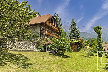 A traditional stone & wood house with balconies overlooks a green yard & trees, set against a mountain backdrop under a blue sky.