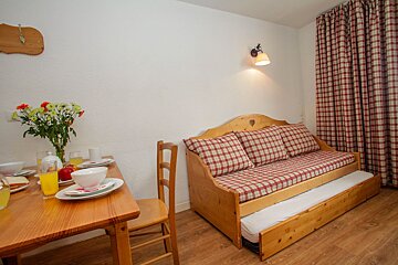 A cozy room with a dining table set for breakfast and a wooden daybed with red plaid cushions and a pull-out mattress, beside matching curtains.