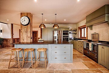 An inviting farmhouse kitchen with sage green cabinets, a central island with stools, warm wood accents, and stone tile flooring.