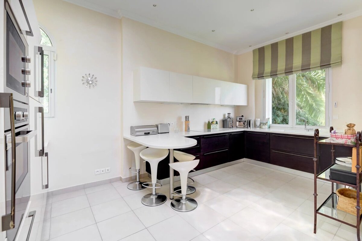 A modern, bright kitchen with white and dark cabinets, tiled floors, a curved breakfast bar with stools, and a large window.