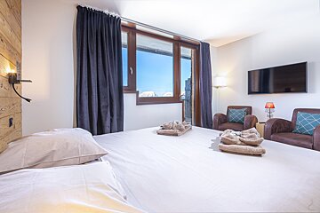 Bright bedroom featuring a white bed with folded towels, dark curtains over a large window, and two armchairs next to a TV.