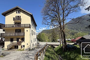 A yellow alpine building with balconies, trees, a road, a small bridge over a stream, and green mountains under a bright blue sky.
