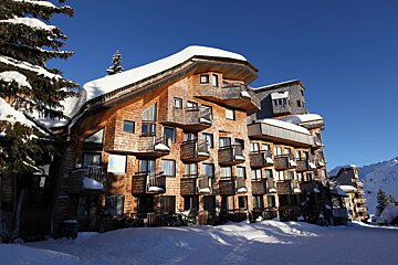 A snowy wooden hotel with many balconies under a clear blue sky, surrounded by snow and a pine tree. Winter mountain scene.