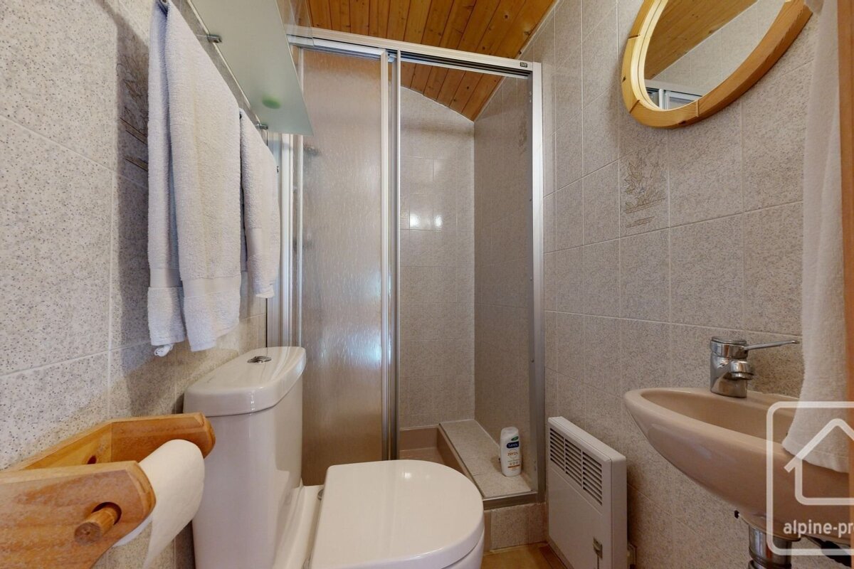 A compact bathroom featuring a toilet, glass-enclosed shower, sink, tiled walls, and a warm wooden ceiling.