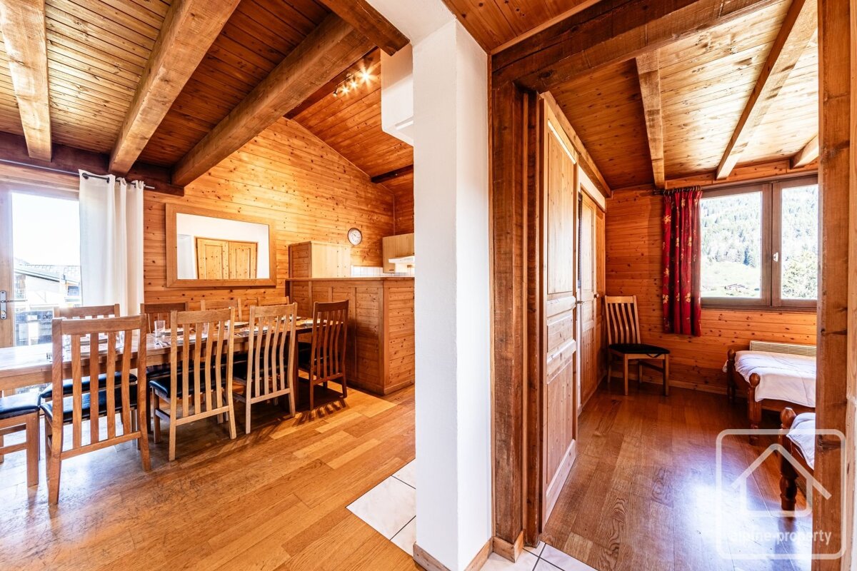 Warm, rustic wooden chalet interior featuring a dining area, kitchen, and a bedroom with a window overlooking mountains.