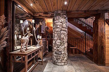 Rustic wood-paneled room with a stone pillar, taxidermy heads, deer statue on a console, and a wooden staircase.