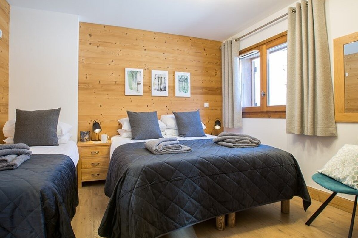 Cozy cabin-style bedroom featuring two beds with dark grey quilted bedding, wood-paneled walls, and a window with curtains.