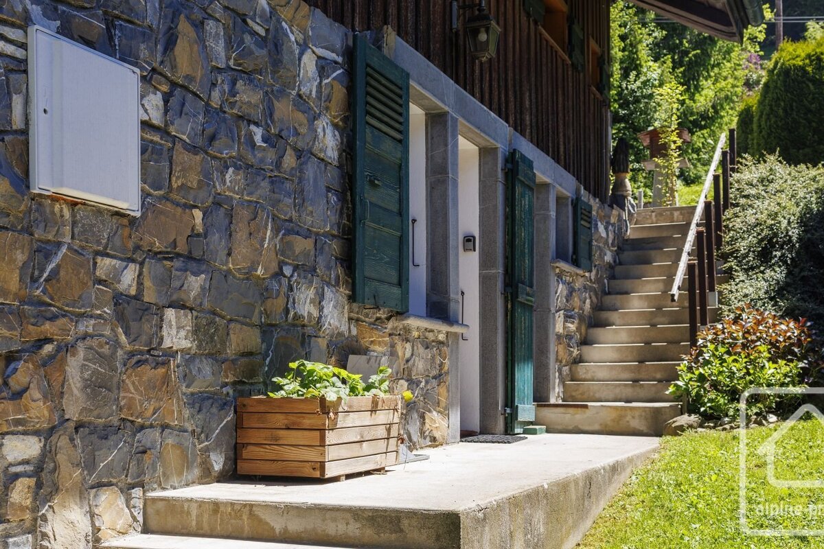 A stone-walled building with dark green shutters features a wooden planter, a concrete path, and sunlit stairs leading up a grassy hillside.