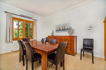 A bright dining room features a wooden table with dark chairs, a sideboard, and a window overlooking greenery. A sailboat painting decorates the white wall.