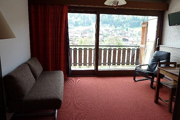 Cozy living room with a brown sofa, red carpet, and large sliding doors opening to a balcony with mountain village views.
