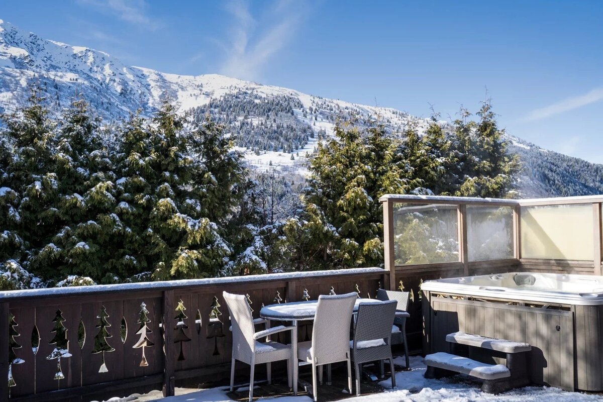 Snowy patio with a hot tub and dining set, overlooking evergreen trees and a majestic snow-covered mountain under a bright blue sky.
