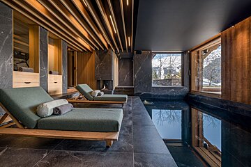 Modern indoor pool with dark marble, wood-slat ceiling, comfy loungers, fireplace, and expansive windows showcasing snowy mountain scenery.