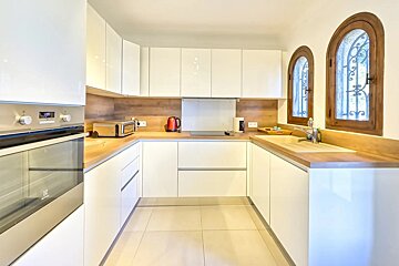 A bright, modern U-shaped kitchen with glossy white cabinets, wooden countertops, and two arched windows with decorative grilles.