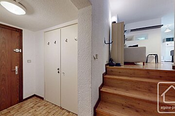 A hallway with a wooden door, white closets with hooks, and wooden stairs leading up to a bright living area/kitchen.