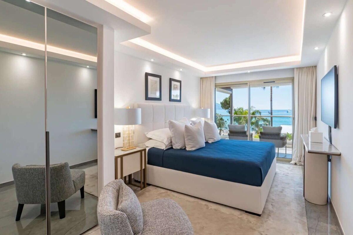 A bright, modern bedroom featuring a blue bed, contemporary decor, mirrored closet, and a balcony with stunning ocean and palm tree views.