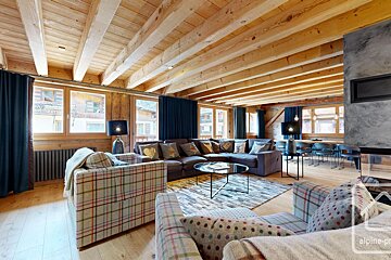 A cozy, spacious chalet living room with a beamed wooden ceiling, plush L-shaped sofa, plaid armchairs, a modern fireplace, and large windows.
