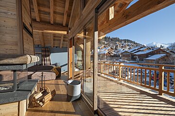 Cozy wooden chalet interior with a fireplace, large windows, and a sunny balcony overlooking a snow-covered mountain village.