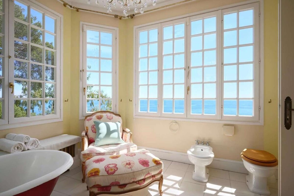 Sunny bathroom with large windows overlooking the ocean and trees. Features a decorative armchair, bidet, toilet, and red tub.