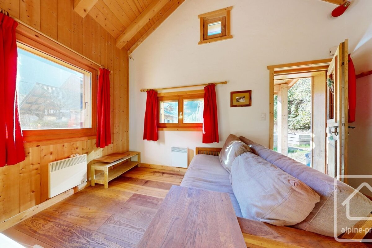 A bright, rustic room with wood-paneled walls, a wooden floor, and a couch. Red curtains adorn multiple windows, and an open door leads to the outdoors.
