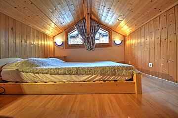 A warm, all-wood attic bedroom with a bed, fluffy blanket, and a central window framed by blue curtains. Wall sconces provide cozy lighting.