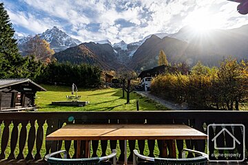 Sunny alpine valley view from a balcony table. Snow-capped mountains, green fields, and houses are visible under a bright, cloudy sky.