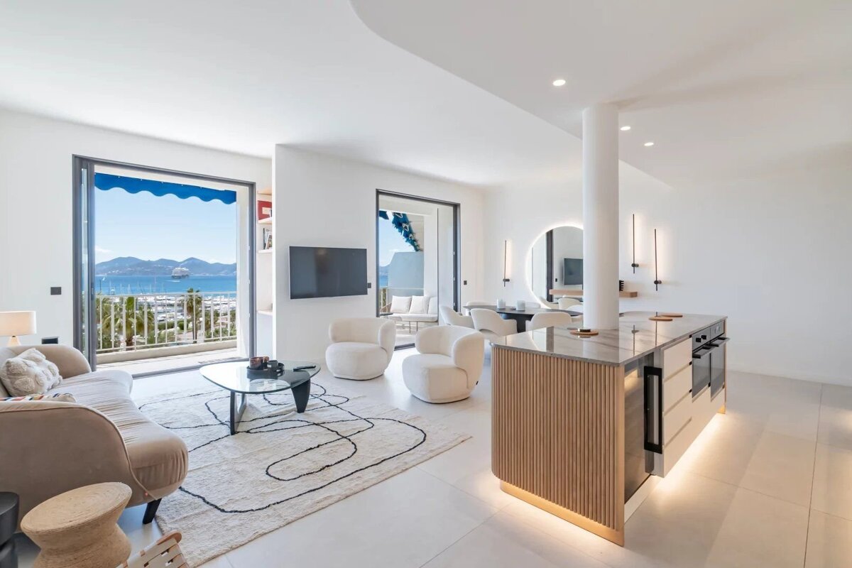 Modern, bright open-plan living area and kitchen with elegant minimalist decor, featuring large doors that open to a scenic ocean view.