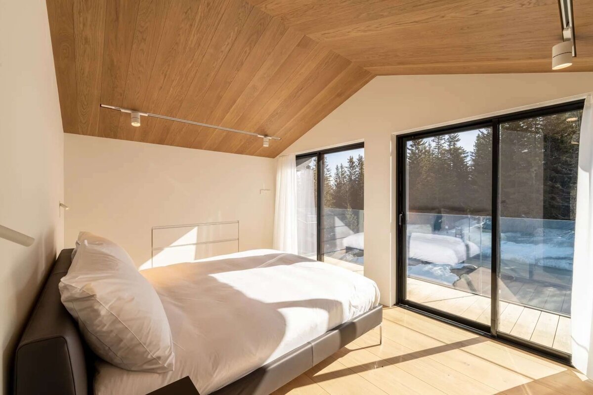 A modern, sunlit bedroom with a comfy bed, vaulted wooden ceiling, and light wood floor. Large glass doors open to a balcony with a snowy forest view.