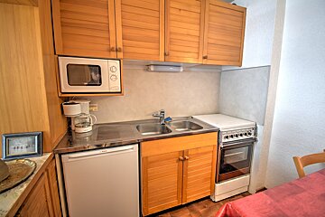A compact kitchen with light wood slatted cabinets, a double sink, a mini-fridge, a microwave, and a small stove.