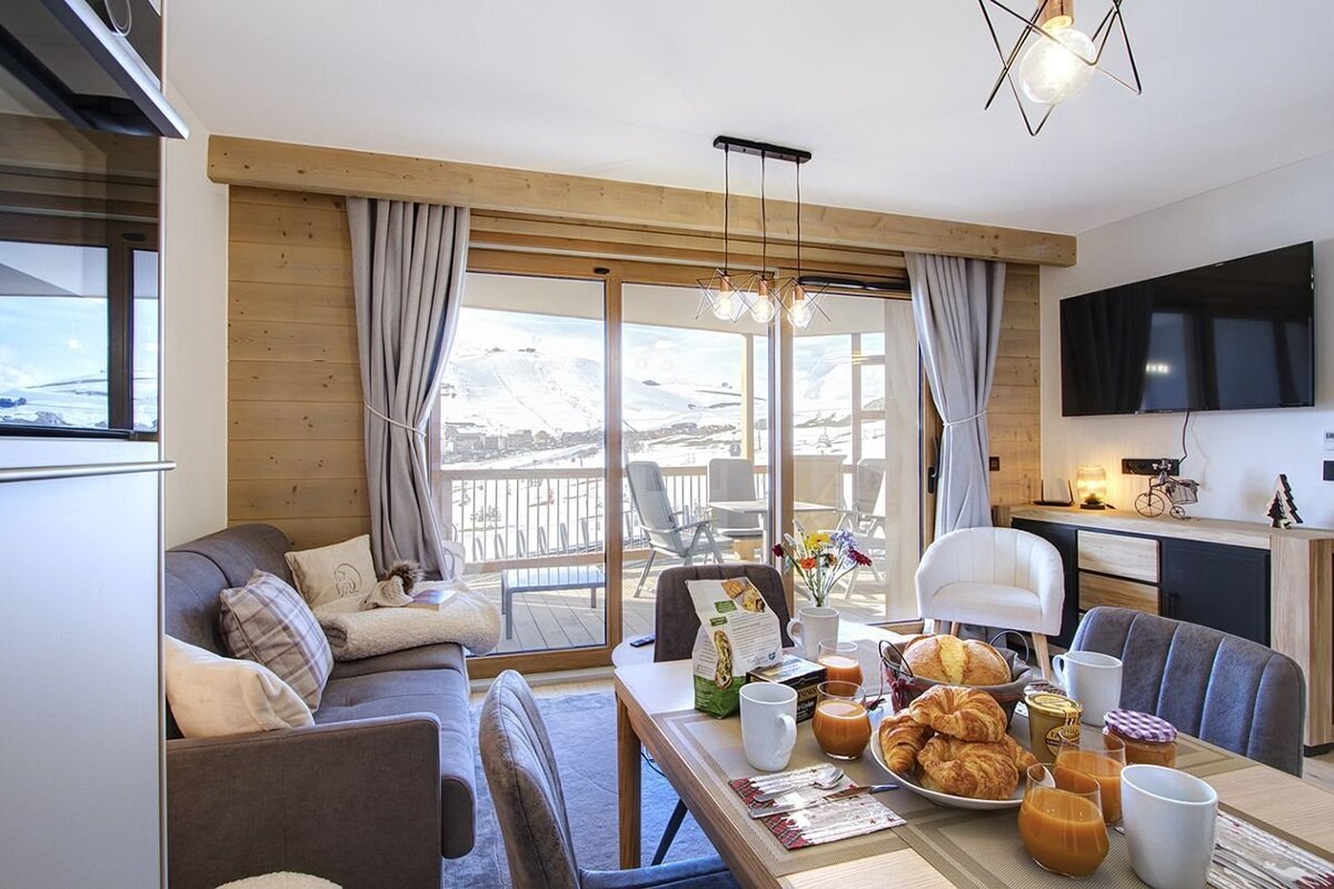 A cozy, modern living/dining room with a breakfast spread, overlooking a snowy mountain landscape from a balcony.