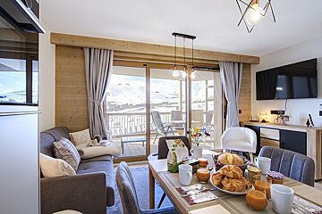 A cozy, modern living/dining room with a breakfast spread, overlooking a snowy mountain landscape from a balcony.