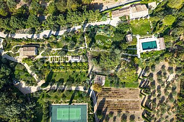 Aerial view of a sprawling estate with houses, lush gardens, a pool, tennis court, and olive orchards amidst green foliage.
