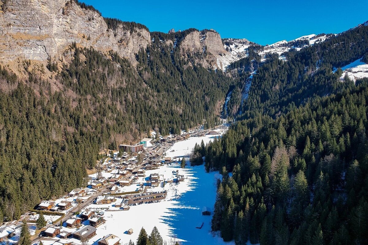 Aerial view of a snowy mountain village nestled in a valley, flanked by steep, forested peaks under a clear blue sky.