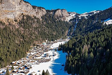 A snowy mountain village nestled in a valley, flanked by dense pine forests and towering rocky peaks under a clear blue sky.