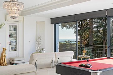 A luxurious living room with a red pool table, white sofas, a crystal chandelier, and large glass doors offering a beautiful sea view.