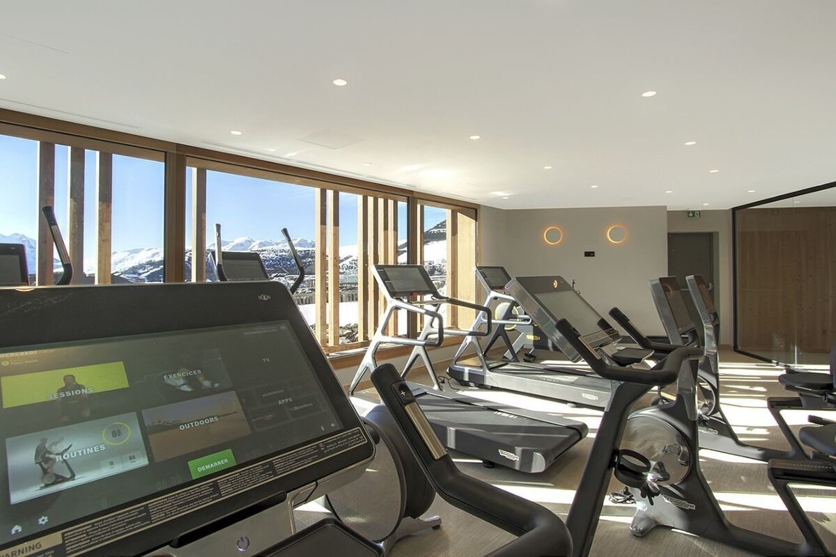 A modern gym with multiple exercise machines, featuring large windows overlooking a stunning snowy mountain landscape.